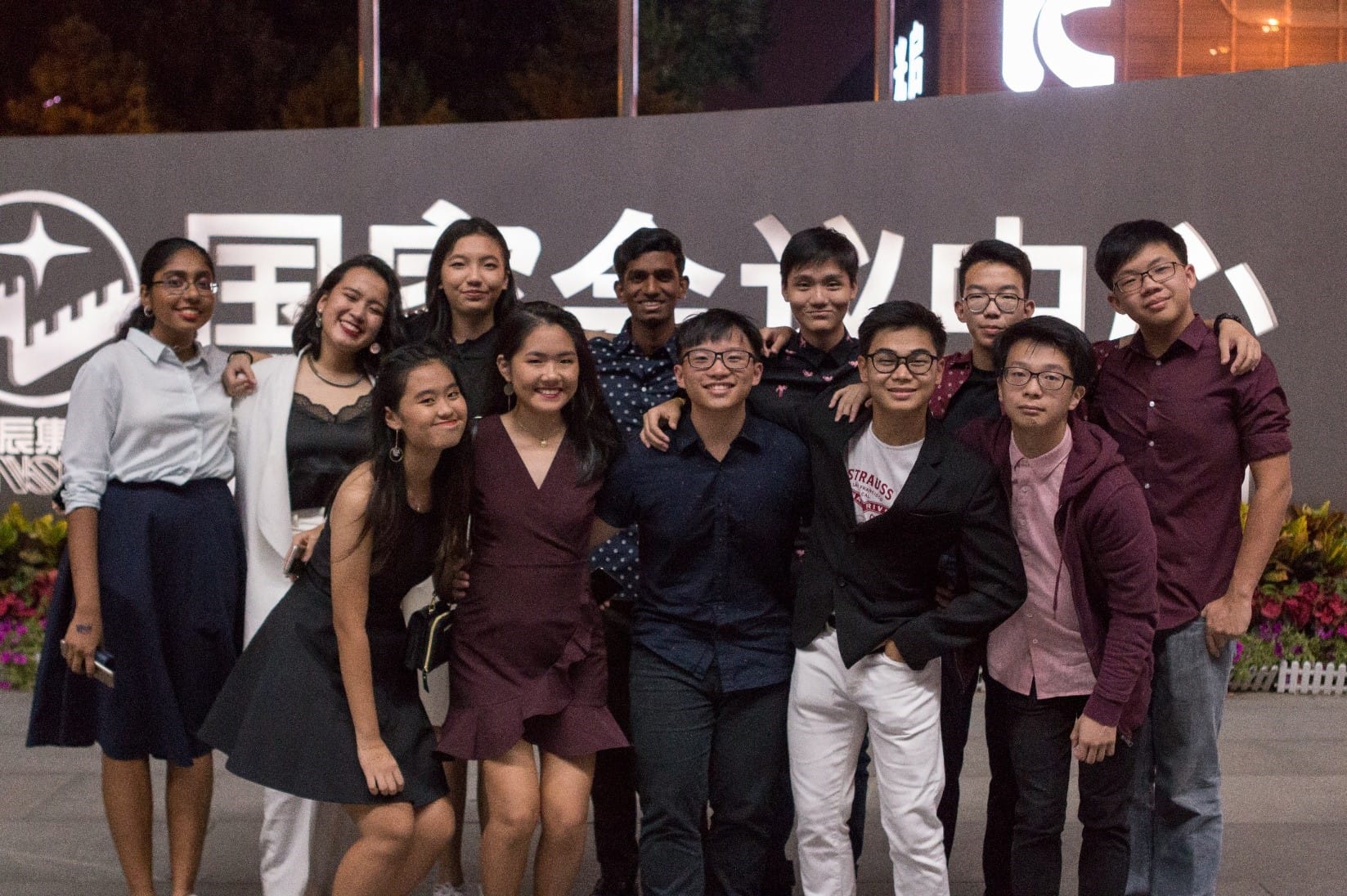 A group of about twelve smiling teenagers posing together outdoors at night in front of a large sign with Chinese characters.